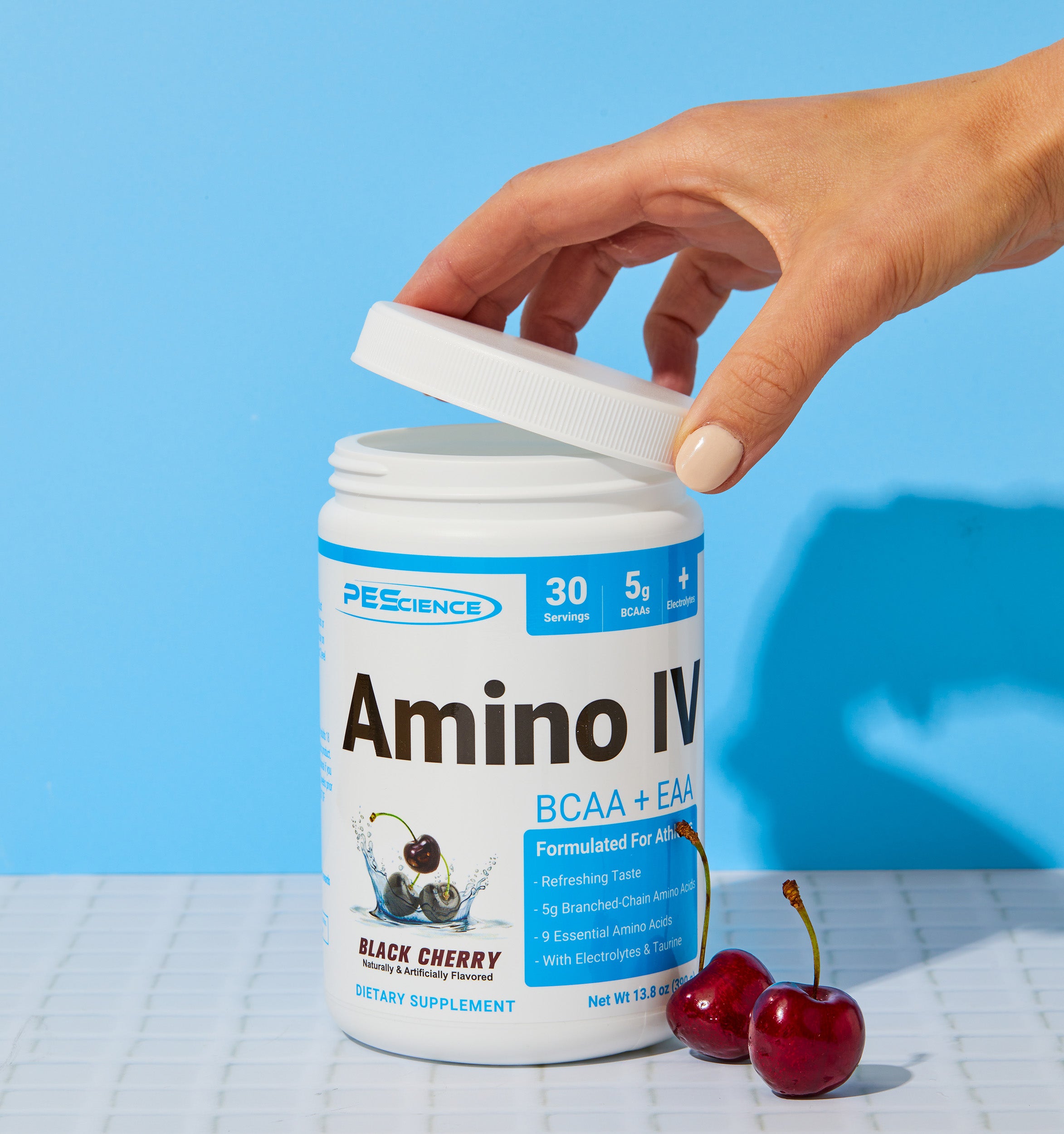 A hand opens a container of PEScience Amino IV dietary supplement in black cherry flavor, with two fresh cherries and the container label visible on a white tile surface against a blue background.