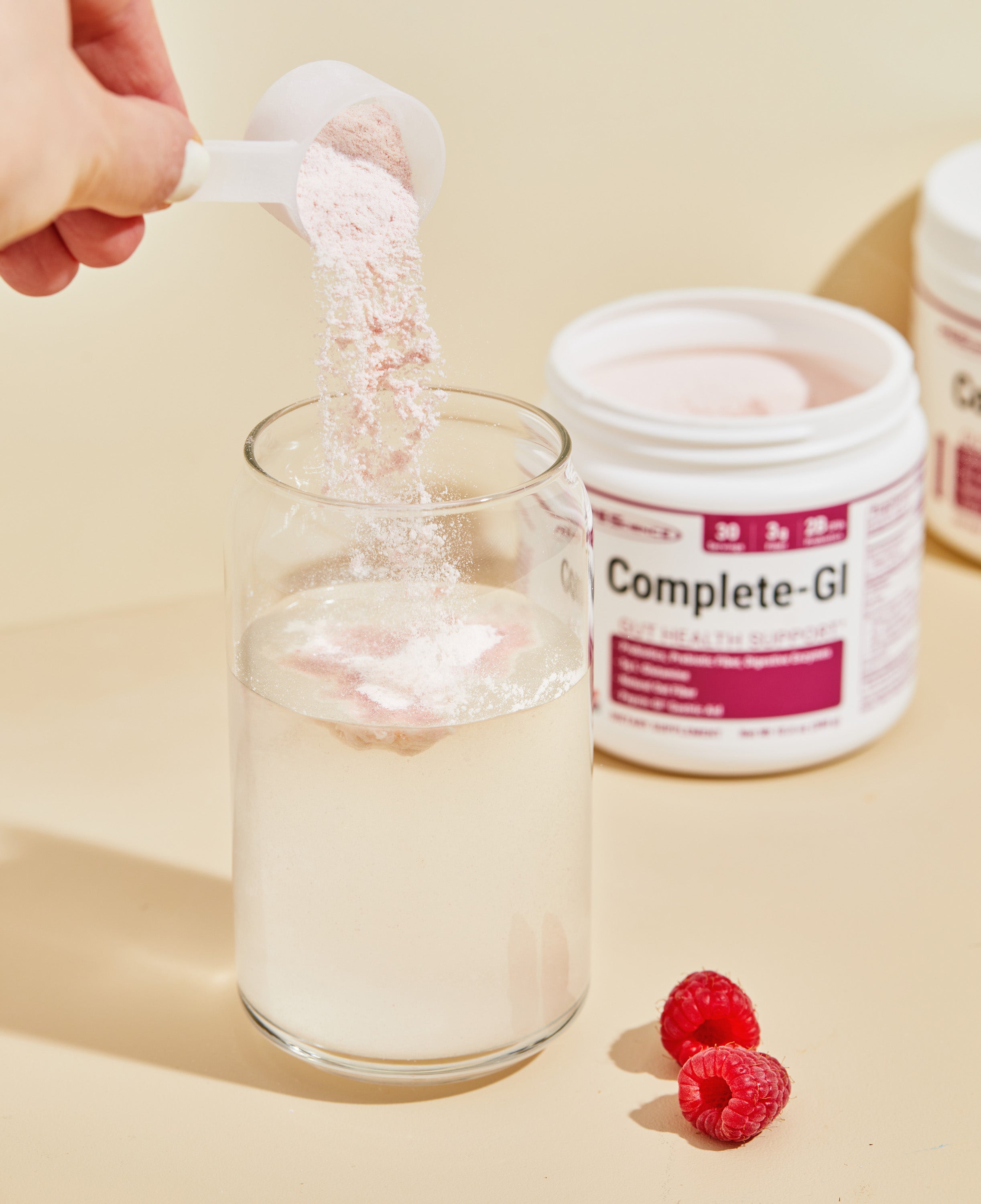 A hand pours a scoop of pink powder into a glass of water. An open container labeled Complete-GI and two raspberries are nearby on a beige surface.