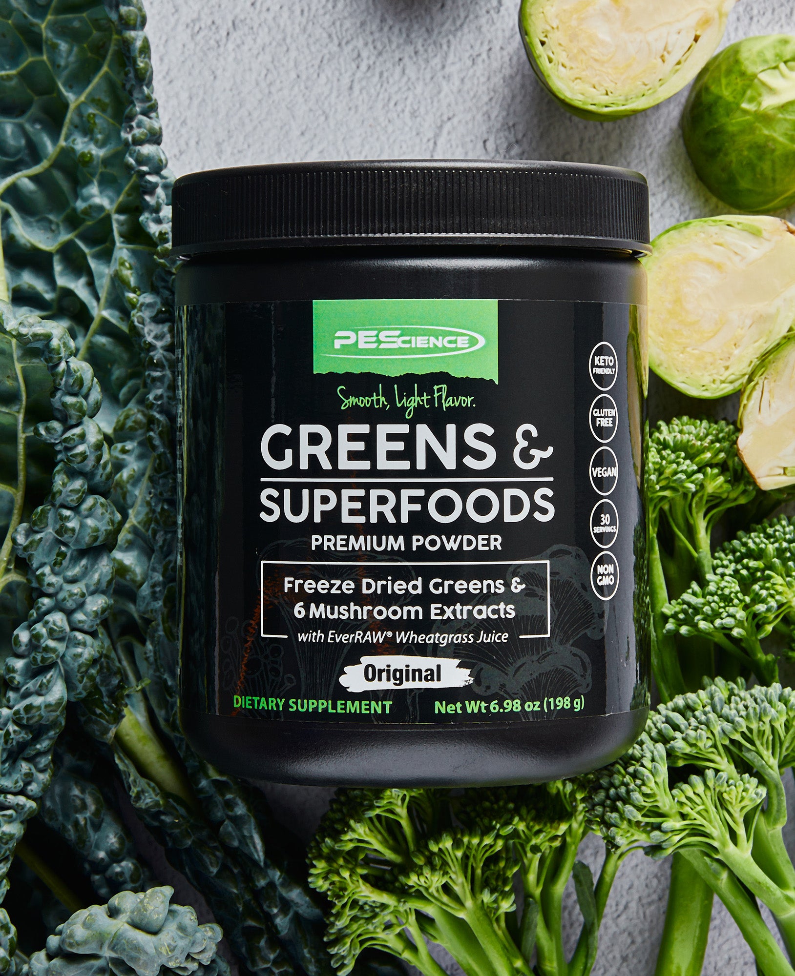 A container of PEScience Greens & Superfoods powder sits on a gray surface surrounded by fresh kale, broccoli, and halved Brussels sprouts. The black container features green and white labeling emphasizing its natural ingredients.