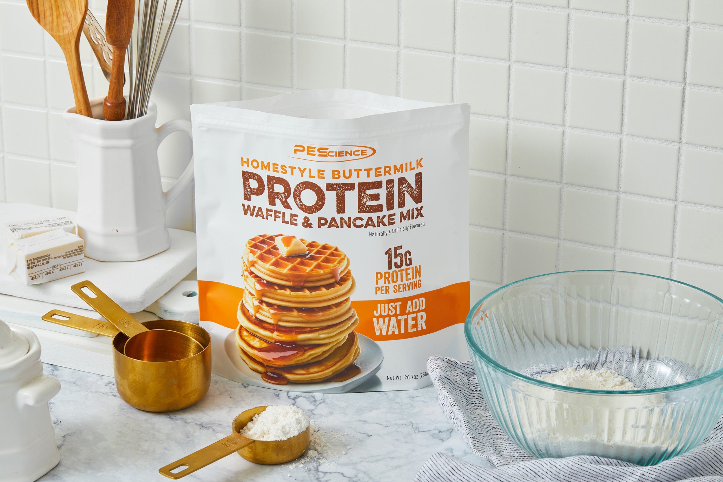 A bag of PEScience Homestyle Buttermilk Protein Waffle & Pancake Mix stands on a kitchen counter next to measuring cups, a bowl with mix, a striped towel, and kitchen utensils against a white tiled backsplash.