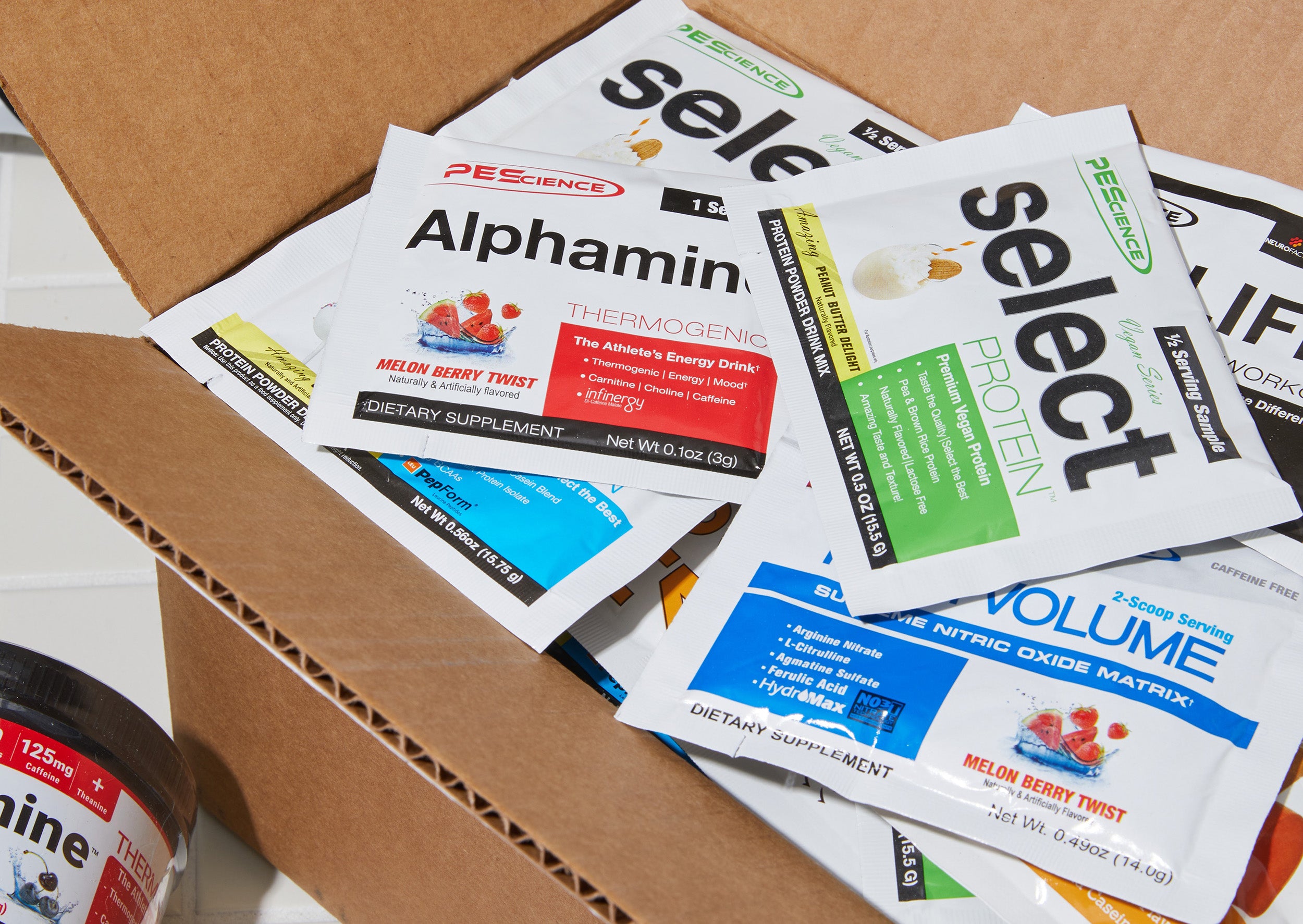 Packets of PEScience dietary supplements, including Alphamine, Select Protein, and High Volume, are spread on and around a cardboard box. The flavors listed are melon berry twist and gourmet vanilla.