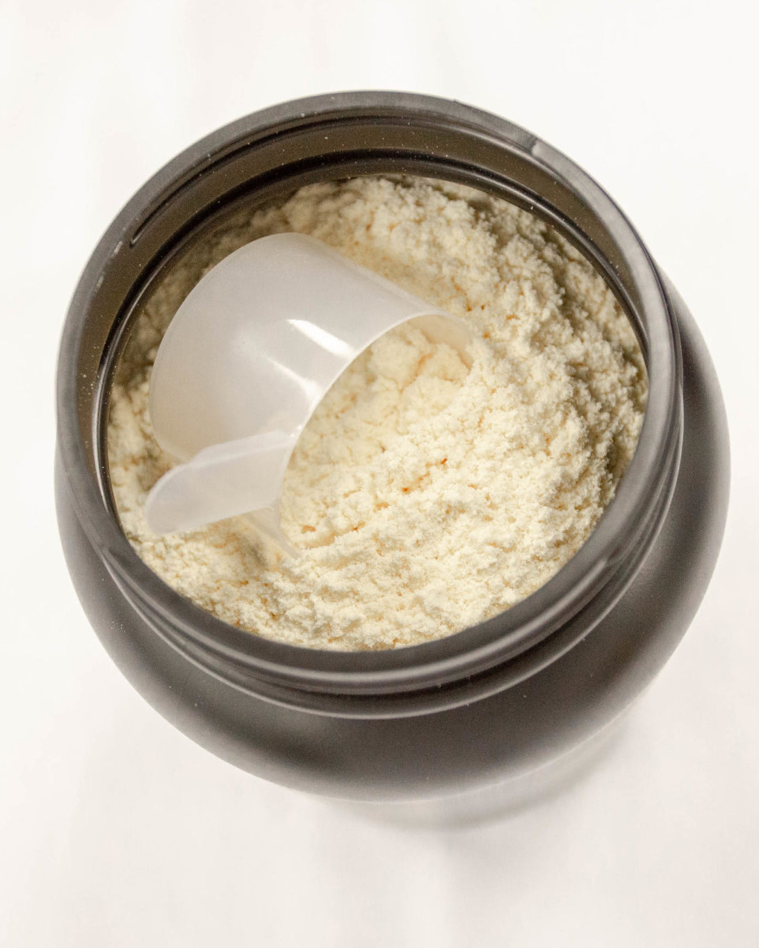 An open black container filled with light-colored protein powder, with a transparent plastic scoop resting on top of the powder.